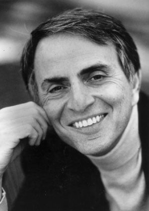 Carl Sagan in his classic turtleneck under a dark blazer, gently resting his right hand on his cheek and smiling, in a black and white photo.