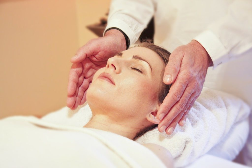 Reiki practitioner holding their hands next to a female-presenting person's head with their fingers pointing towards her shoulders. She is lying on their back facing up, with their head supported on a towel and with a towel covering their body. The Reiki practitioner standing behind her appears to be a man and he is wearing a white outfit.