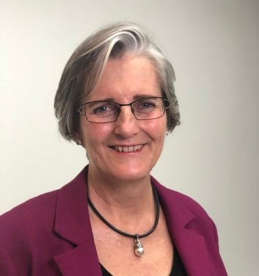 Dr Joy Bowles. She is smiling and has a cool short hair cut with highlights of silvery grey through the front. She is wearing metal framed glasses, a chunky black necklace and a dark magenta blazer. Image source: https://www.ejoybowles.com/bio.html#/