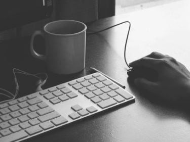 Keyboard and mouse with headphones and coffee cup behind. A hand is on the mouse. 