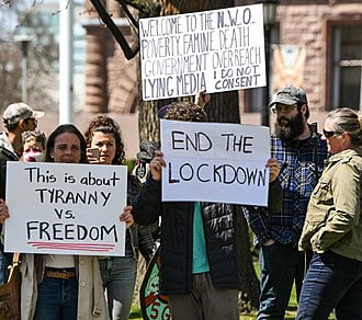 People protesting at Queen's Park, Toronto in April 2020 - signs read "End the lockdown", "welcome to the NWO, poverty, famine, death, government over reach, lying media, I do not consent" and "this is about tyranny vs. freedom".