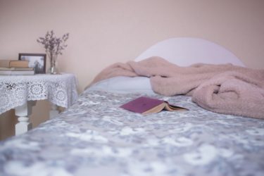 A bed in a soft pink room with grey sheets and a white table beside it. A pink book lays open on the bed.