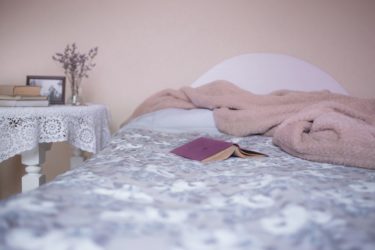 A bed in a soft pink room with grey sheets and a white table beside it. A pink book lays open on the bed. 