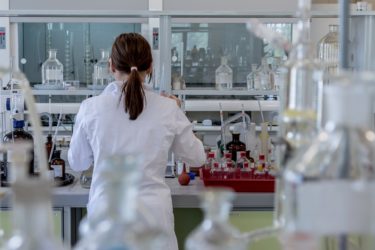 A woman from behind as she stands in a lab wearing a lab coat