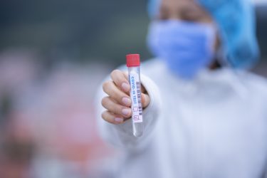 A person wearing a lab coat, face mask and hairnet holds out a sample tube with the words "COVID-19 TEST" alongside it. 
