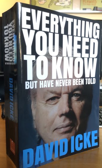 David Icke's book: Everything you need to know but have never been told. The image on the cover is a close up of his face. The title is in large white block capitals across the top and his name in blue block capitals across the bottom. The book is standing on a desk in front of a computer. 