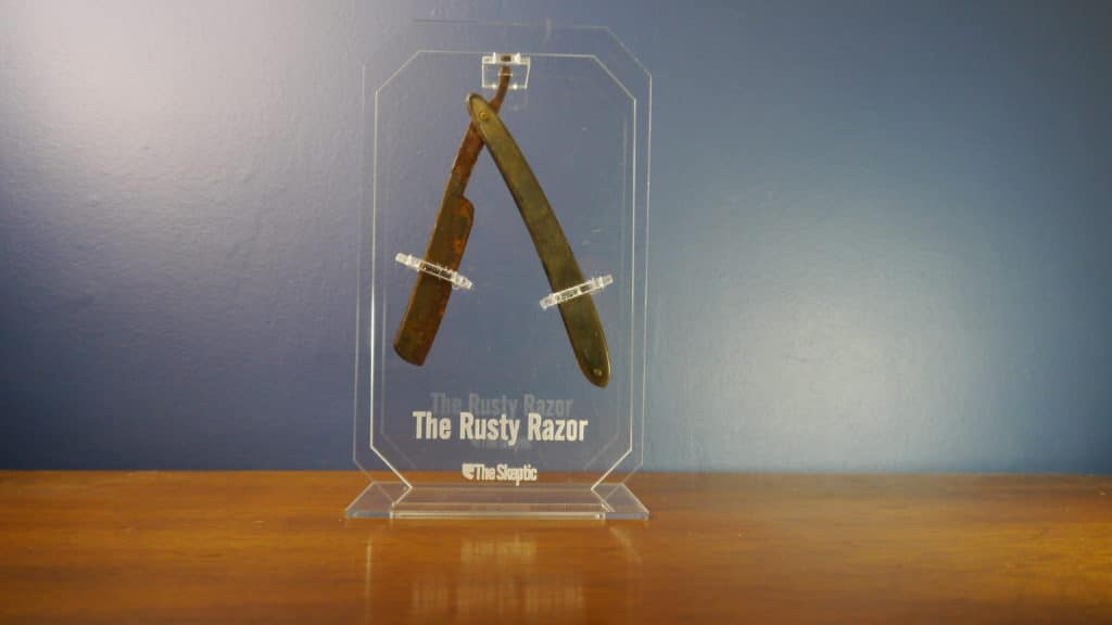 A photograph of the Rusty Razor award: a rusty classic style straight razor encased in plastic with "The Rusty Razor" etched at the bottom. 