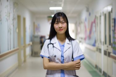 A health care worker standing in the corridor of a hospital. She is wearing a lab coat and a stethoscope and blue scrubs.