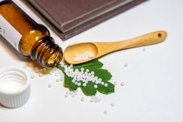 A bottle of homeopathy, open with pills spread across a leaf and onto a table top. There is a wooden spoon and a book beside the bottle and pills.