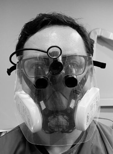 Shaun is wearing a high filtration face mask and visor. The face mask includes two large filter packs on either side and straps that cross the cheeks, over the ears and around the back of the head. Between his regular glasses and the visor are a pair of dental magnifying loupes and above, a small head torch. The only part of Shaun's skin that is clearly visible is his forehead.