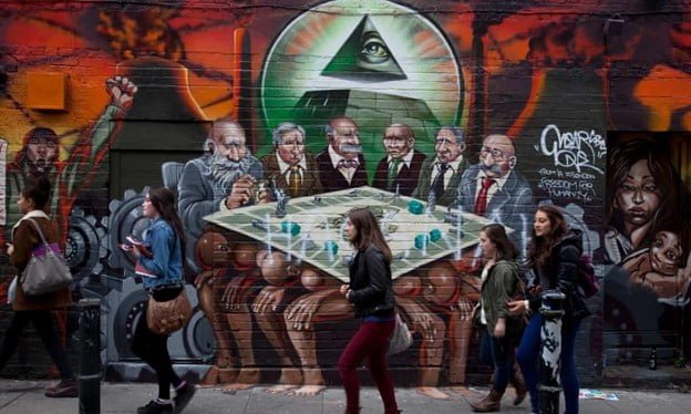 A group of Jewish or Eastern European looking men who are meant to appear as bankers sit at a Monopoly board that is resting on the backs of four muscular men of color who are sitting bent over with their heads between their knees. On the Monopoly board are several small pewter pieces like a boat and a statue of liberty and some green Monopoly houses. Behind the bankers is an Illuminati Eye of Providence pyramid flanked by power plants, giant gears, and both refuges and revolutionaries of color. The mural has been defaced with the word “Haganah”, referring to a defunct Jewish paramilitary organisation