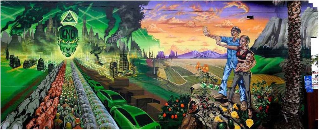 On the left half of the mural is a dystopian landscape with lines of animals, humans, and machines all converging towards a smoking skyline centered around a giant floating head with “GMO” printed on the forehead and an Eye of Providence floating over the heat. On the right side of the mural is a native family, man woman and baby, standing in a verdant utopia with their arms stretched out in a stop sign