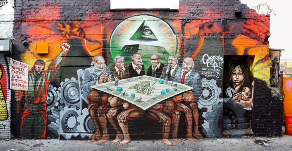 A mural featuring a group of white men who are meant to appear as bankers sat at a Monopoly board that is resting on the backs of four muscular men of colour, who are sitting bent over with their heads between their knees. On the Monopoly board are several small pewter pieces like a boat and a statue of liberty and some green Monopoly houses. Behind the bankers is an Illuminati Eye of Providence pyramid flanked by power plants, giant gears, and both refuges and revolutionaries of colour. The mural has been defaced with the word “Haganah”, referring to a defunct Jewish paramilitary organisation