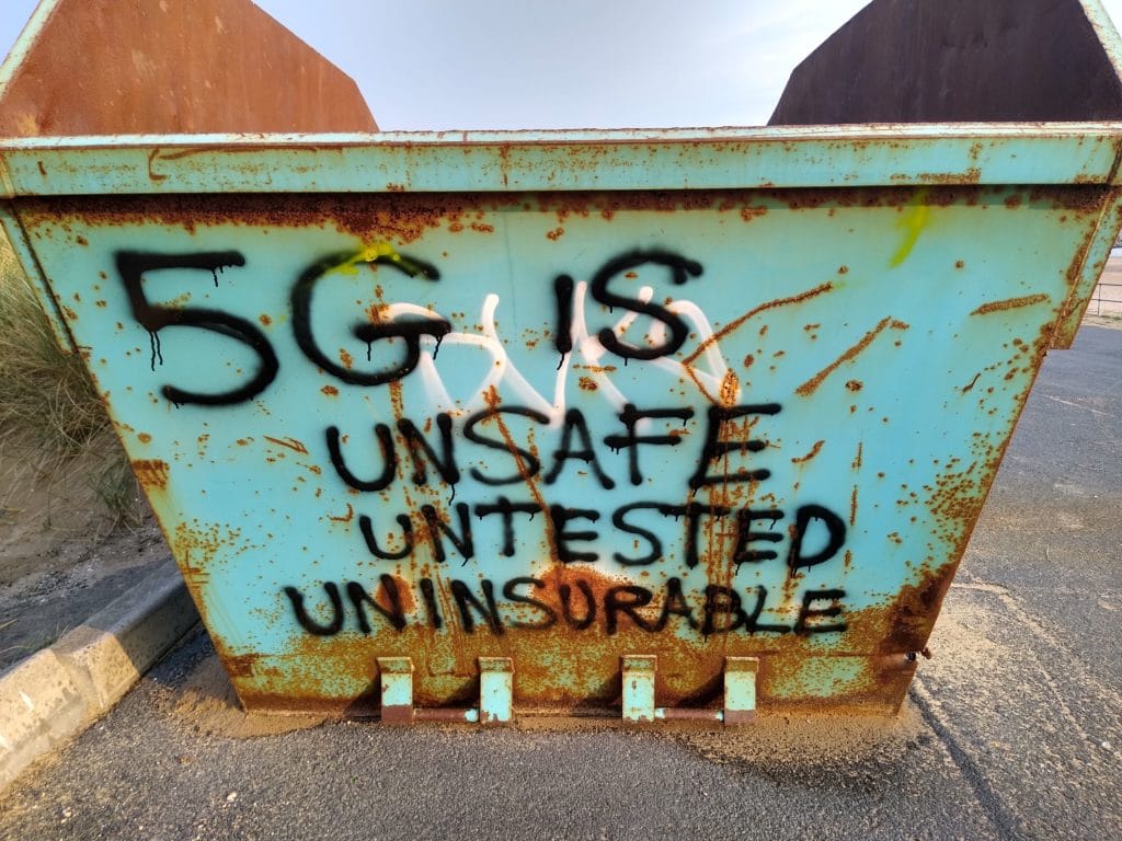 A green, rusted skip spray painted with the words "5G is unsafe untested uninsurable" in black paint. 