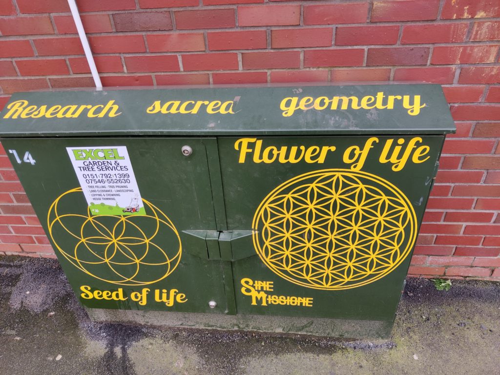 A metal box with Sine Missione artwork on it - the words "research sacred geometry", "flower of life", "seed of life" and two geometric patterns are seen in yellow. One of the patterns is partially covered by an unrelated advert for gardening services. 