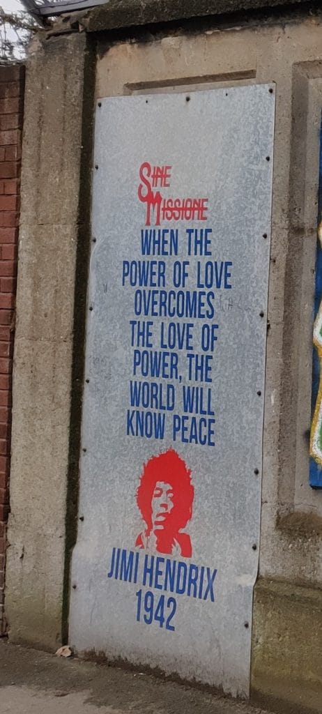A piece of typical sticker art from Sine Missione on a metal board in Liverpool. The artist's name is written in red at the top, below that the words "when the power of love overcomes the love of power, the world will know peace" in blue. Below that an image of Jimi Hendrix, stencil style, in red and the words "Jimi Hendrix, 1942" below in blue.