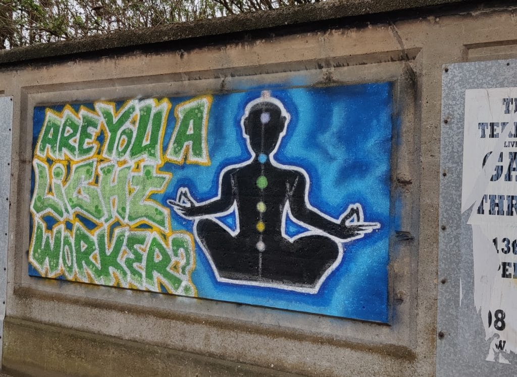 A wall with a rectangle spray painted in blue. On top is a black silhouette sitting in a seated yoga pose (sukhasana) with the seven chakra painted on top. Next to this figure are the words "Are you a light worker?" in large green graffiti style writing.  