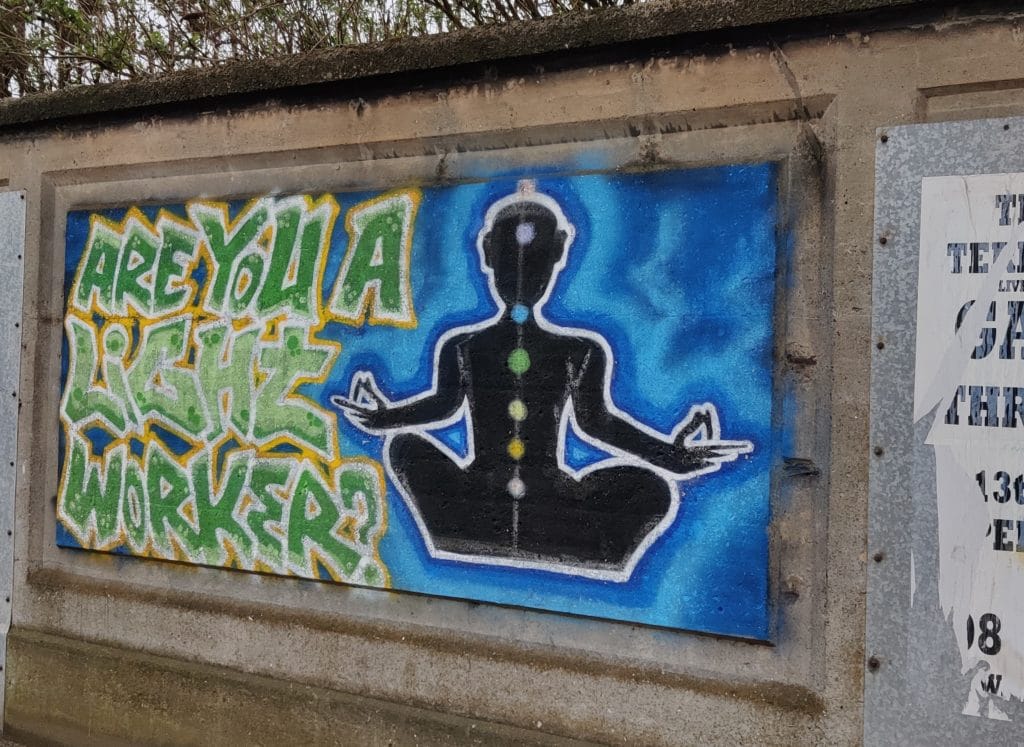 A wall with a rectangle spray painted in blue. On top is a black silhouette sitting in a seated yoga pose (sukhasana) with the seven chakra painted on top. Next to this figure are the words "Are you a light worker?" in large green graffiti style writing.