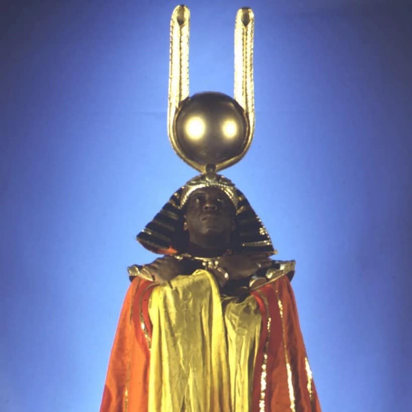 An image of Sun Ra. He is a black man wearing a red and yellow gown with gold detailing. The shoulders have large shoulder pads with gold detailing. On his head, Sun Ra wears an ancient Egyptian style headdress with a large gold ball on the top of it similar to that of the ancient Egyptian god, Ra. He is looking up to the sun shaped ball and has his arms across his chest.  