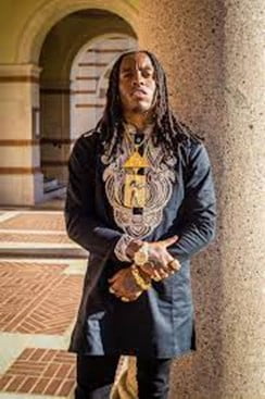 Young Pharoah - a young Black man with long hair in dreadlock braids. He is standing in a corridor with arches behind him and is wearing a black tunic with gold embroidery across the chest and black jeans. He is wearing a large gold necklace which has a gold pyramid hanging above a square gold plate with a depiction of an ancient Egyptian person on it. He is also wearing a large gold watch, large gold bracelet and large gold rings on each hand.