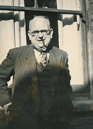 Harold ‘Chib’ Chibbett (1900-1978) - a black and white photo of a balding man with whisps of white hair, dark rimmed glasses and a pipe. He's standing in front of a window, outside, with his hands on the window ledge and he's wearing a three piece suit with a shirt and tie. 