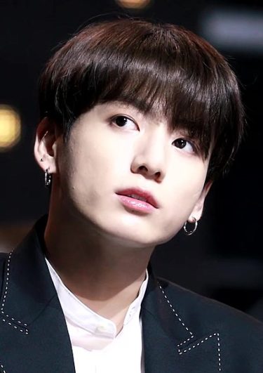 Jungkook, a young South Korean man who is looking up to the corner. He is wearing hoop earrings, a white shirt and a black blazer. 