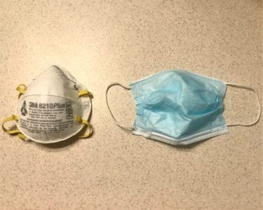 An N95 mask from 3M beside a surgical mask. The surgical mask is a blue piece of material with a metal strip across the note. The N95 mask is thicker material that is structured into a cup shape with a flared edge to sit flush against the skin. It has an adjustable metal nose band. Image from Wikimedia user Rickmouser45 (CC BY-SA 4.0)