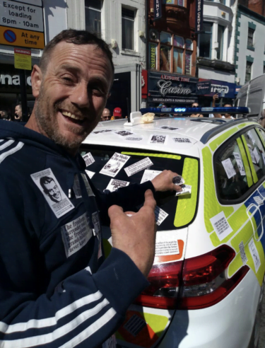A man with a huge smile on his face is standing beside a police car. Both the man and the car are covered in White Rose stickers. The man is pointing at the car with one hand, with the other he is holding a white rose sticker against the car - either removing it, or placing it. In the background, a street in central Liverpool.