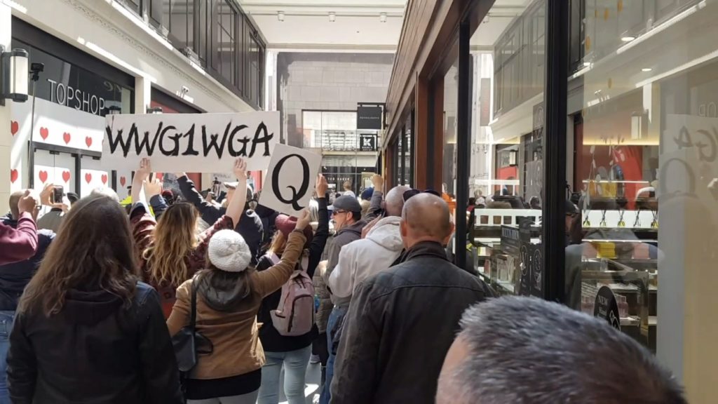 in a covered shopping street in Liverpool city centre a group of people walk - two women hold aloft signs that read "WWG1WGA" and "Q"