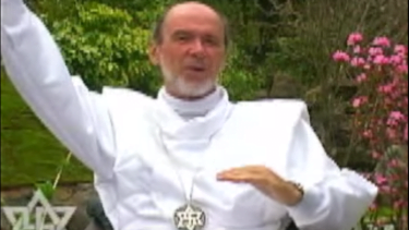 Rael wearing a white outfit with a metal necklace with a Rael symbol