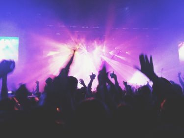 A modern day concert - lights in pinks and purples shine from the stage. The image is taken from the perspective of an audience member looking through a sea of silhouetted hands against the pinky purple light
