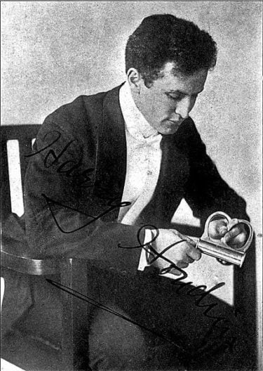 A press photo of Harry Houdini sitting in a chair and looking down at his hands where he holds a pair of handcuffs. He is wearing a black suit with a white shirt and bow tie.