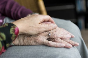 A close up of two pairs of hands. The hands appear to belong to two women, the younger woman's hands are reassuringly held over the top of the older woman's hands. 