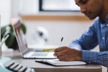 A person of colour who presents as masculine sits at their laptop while writing in a notebook. 