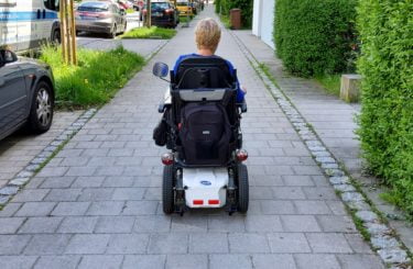 A white woman with a short blonde hairstyle travels down a pavement using her electric wheelchair.