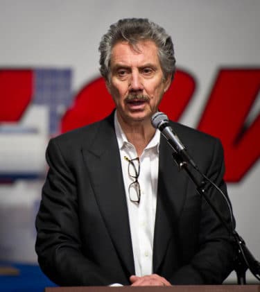 Robert Bigelow standing behind a microphone talking. He is a slim man with grey hair and a grey moustache. He is wearing a crisp white shirt and a black blazer. He has a pair of glasses attached to his shirt. 