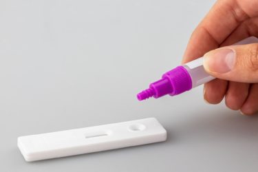 A hand applies a sample from a sample tube on to a COVID-19 lateral flow cassette. 