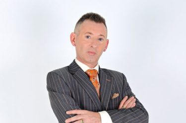 Colin Fry - a thin white man with short grey hair wearing a grey pinstriped suit and an orange tie