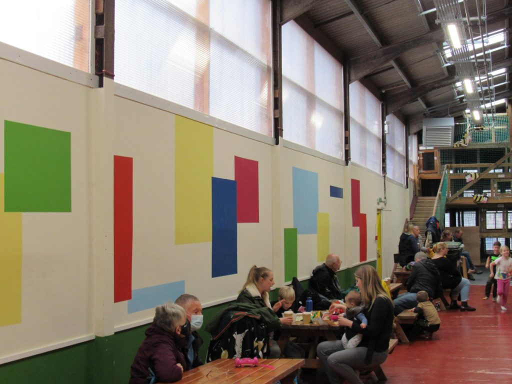 The Ark Arena as seen in October 2021. The walls are devoid of creationist posters, replaced with colourful geometric patterns. There are picnic benches in front of the walls with people sitting. An elevated section of the play area can be seen in the background.