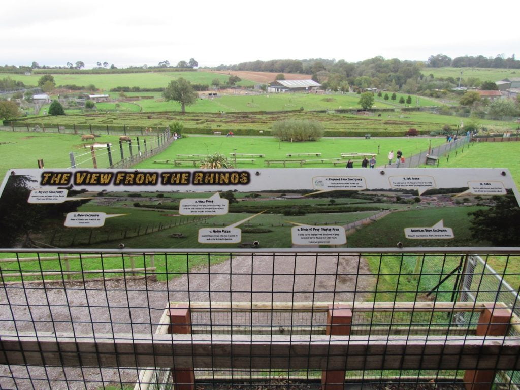 View of the zoo farm taken in October 2021 from a tower by the rhino enclosure. An infoboard is on the barrier highlighting where different areas are. There are enclosures in the foreground, the hedge maze in the mid-ground and Elephant Eden in the background. 