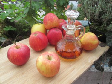 A wooden board stacked with red apples. There is a glass decanter on the board filled with vinegar