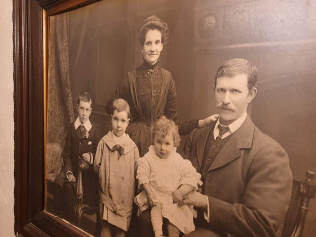 A photo of an old black and white family portrait on the wall. A woman dressed in a black dress with her hair in a bun on top of her head. A man with a moustache and a three piece suit and three young children. 