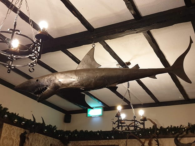 A photograph of a large metal shark hanging from a beamed ceiling. 