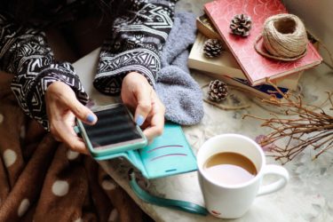 A close up of feminine hands with blue nail polish holding a smartphone in a teal phone case. There are books beside the person and a cup of tea. 