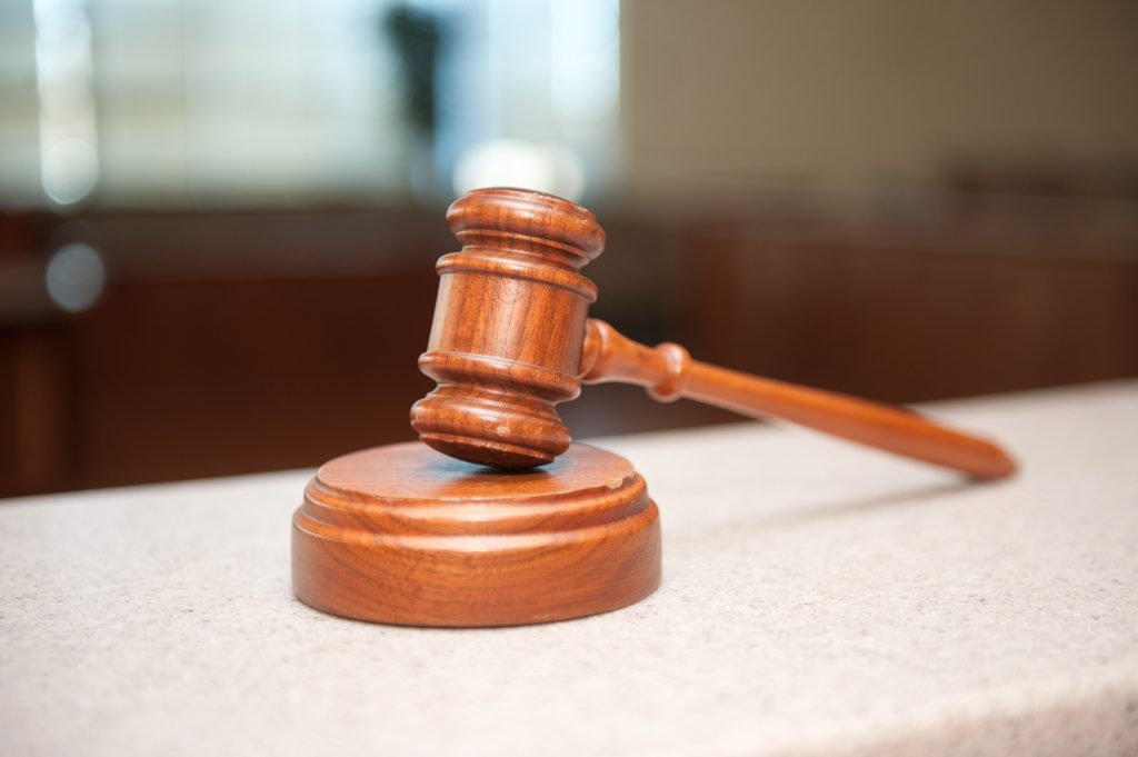 A wooden judge's gavel rests on a pale stone surface in a courtoom, which is blurred in the background