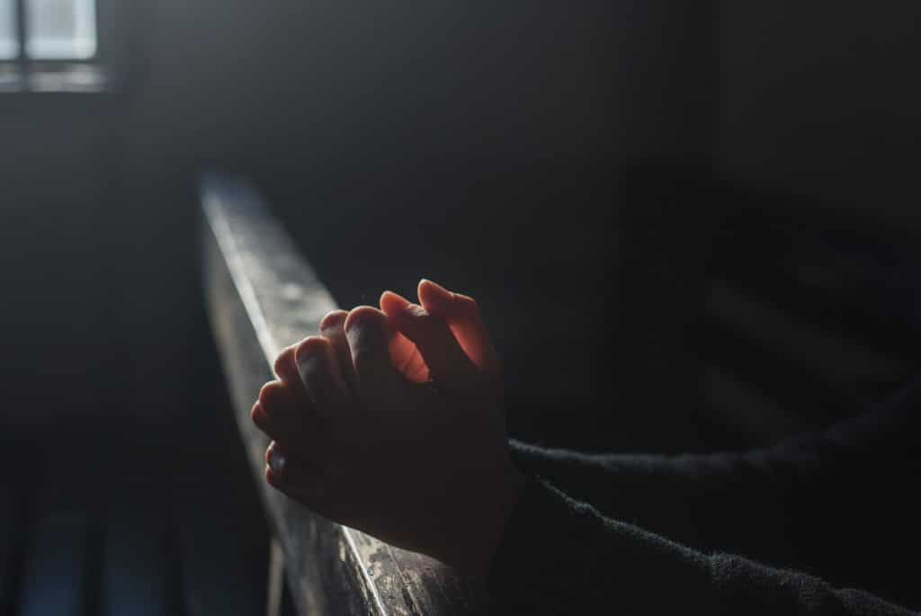 A pair of light-skinned hands clasped in prayer, wrists resting on a wooden railing in low natural light.