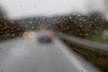 A windshield with rain on it. Beyond the windshield you can see the break lights of the rear of a car in front. 