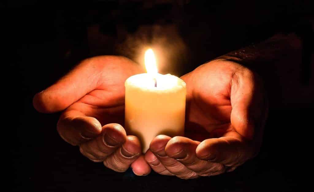 A lit white candle in the palms of a person, the surroundings are pitch black