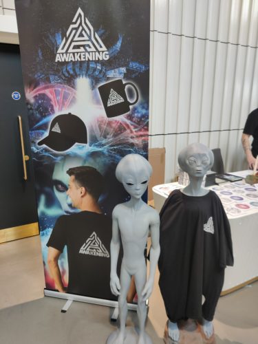 A standing banner for Awakening merch including a baseball cap, a mug and a t shirt. Beside the stand are two grey alien figures, one is wearing the branded t shirt. 