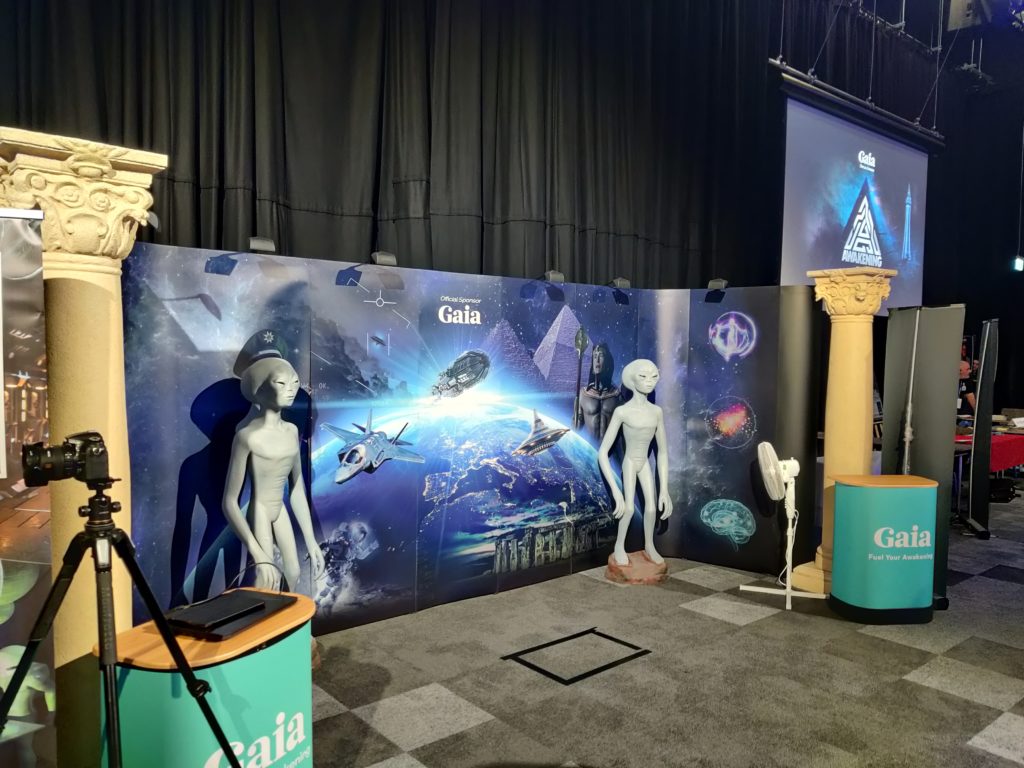 A large advertising banner for Gaia in front of which are two more grey alien figures. The banner has a shot of earth from a distance with various space ships superimposed over the top, and Stonehenge and the pyramids also superimposed in the back- and foreground.